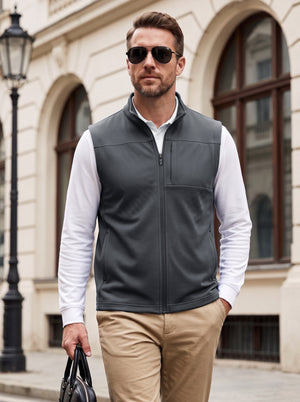 Men's charcoal gray nylon blend golf vest worn over a white long-sleeve shirt with beige trousers, featuring a stand collar, front zipper, textured fabric, and practical pockets in an elegant urban setting.